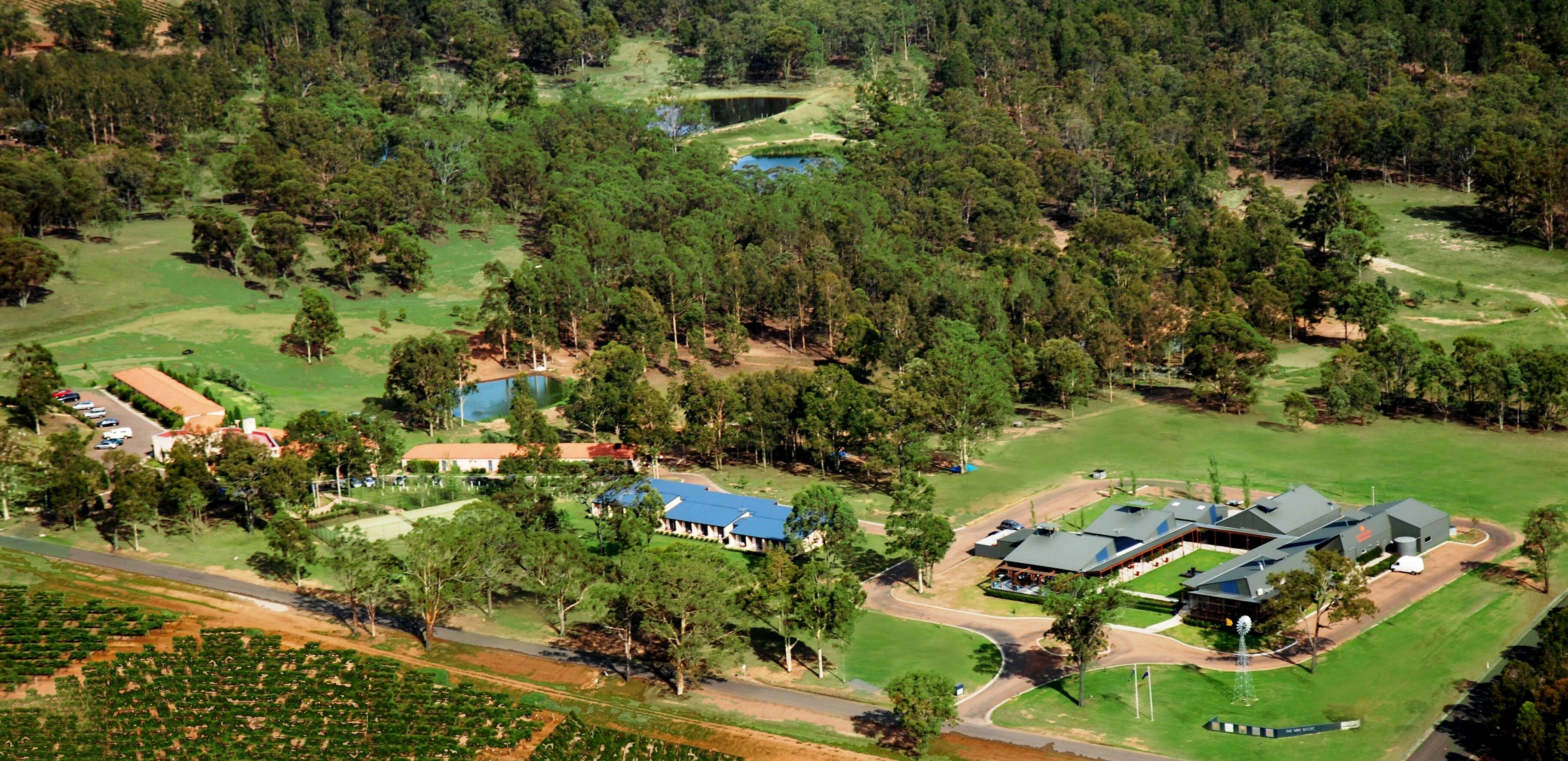 An aerial view of a large estate with multiple buildings, including a main house with a blue roof, a guesthouse, and several smaller structures. There are well-maintained lawns, a golf course, and a pond.