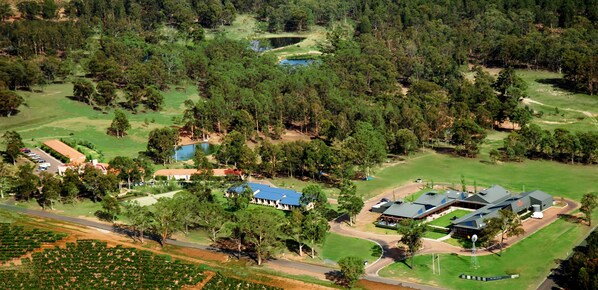 An aerial view of a large estate with multiple buildings, including a main house with a blue roof, a guesthouse, and several smaller structures. There are well-maintained lawns, a golf course, and a pond. - Estate Tuscany (Pokolbin)