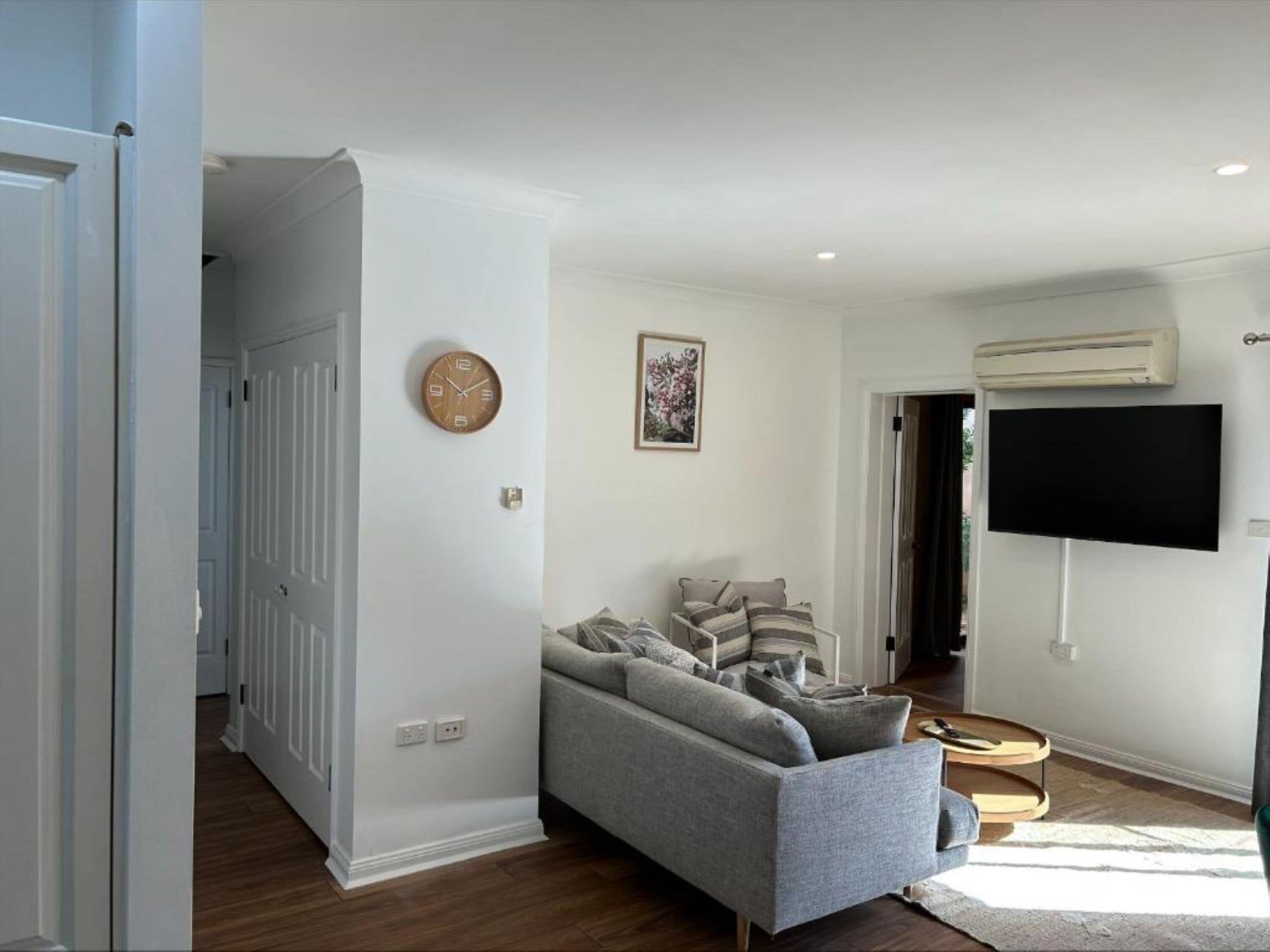 A living room with a grey sofa, a wall clock, and a flat-screen TV mounted on the wall.