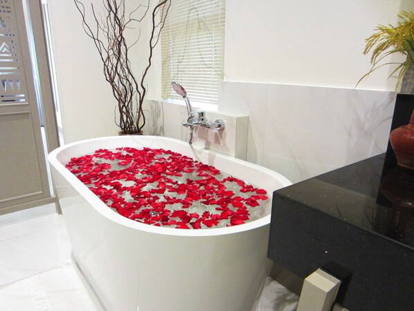 Une salle de bain avec une baignoire sur pieds remplie de pétales de roses, une fenêtre avec des stores et une plante sur la droite.