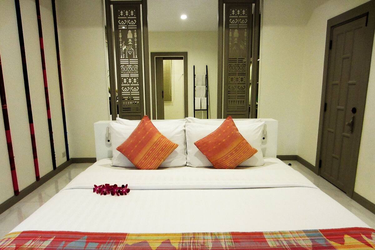 A hotel room with a bed, two pillows, a decorative pillow, a floral arrangement, and a colorful rug.
