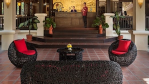 Uma área de recepção com teto de madeira, recepcionista e mobiliário de vime com almofadas vermelhas.