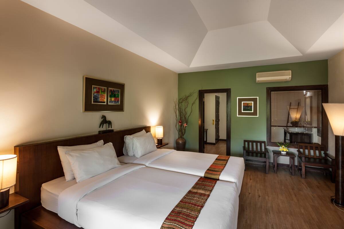 A hotel room with a large bed, two bedside tables with lamps, a small table with a vase of flowers, a wooden bench, and a mirror.
