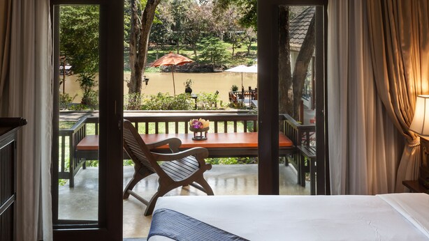 Quarto de hotel com cama, escrivaninha de madeira e vista para área externa com mesa, cadeiras e guarda-sóis.