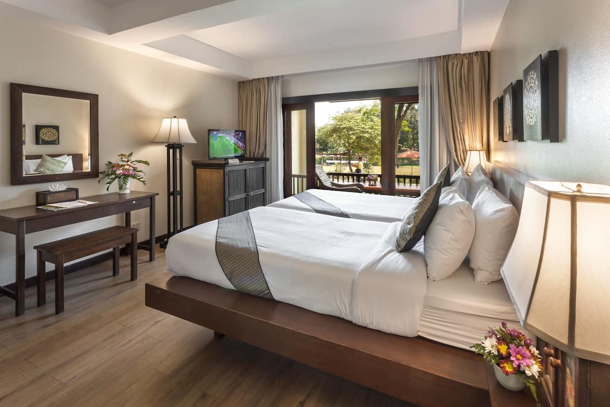 A hotel room with a large bed, a desk, a TV, and a balcony with a view of trees.