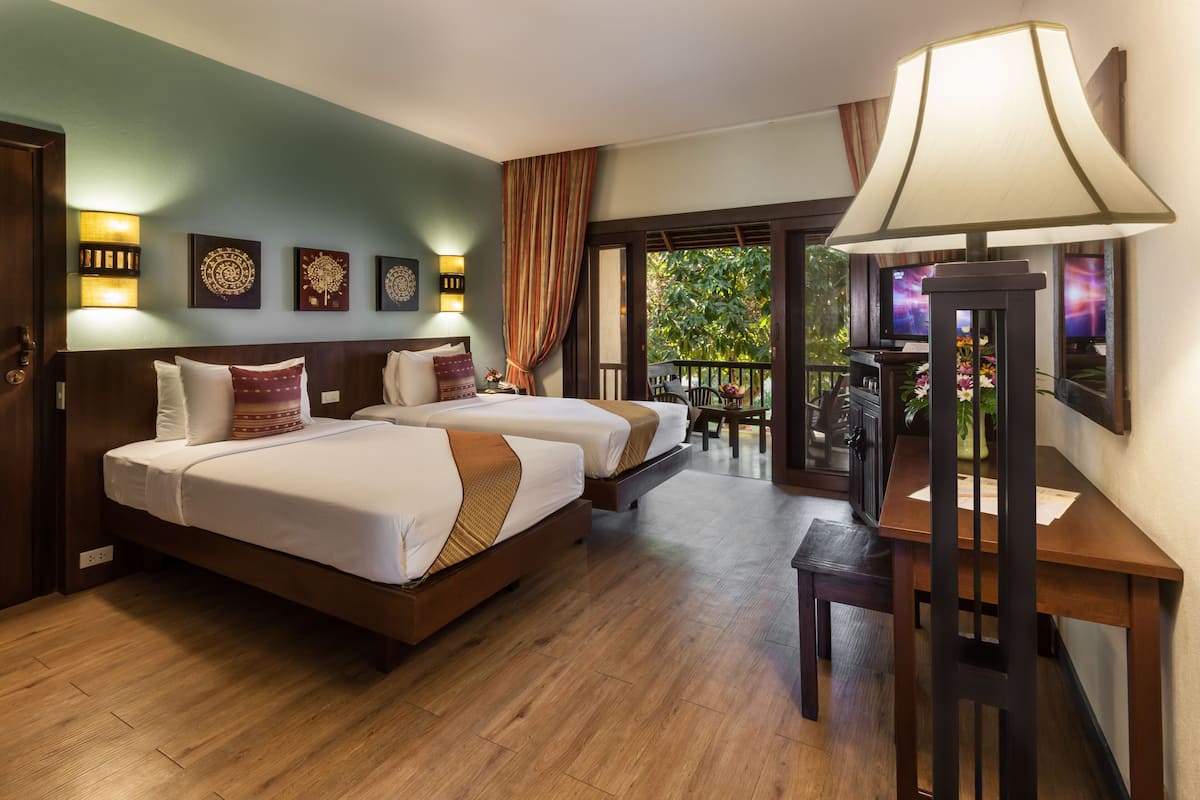 A hotel room with two beds, a desk, a chair, a lamp, and a balcony with a view of greenery.