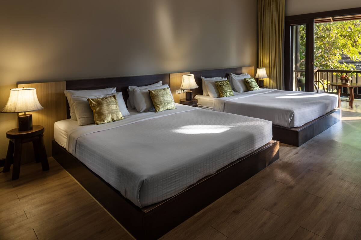 A hotel room with two beds, wooden floors, and a balcony with a view of trees.