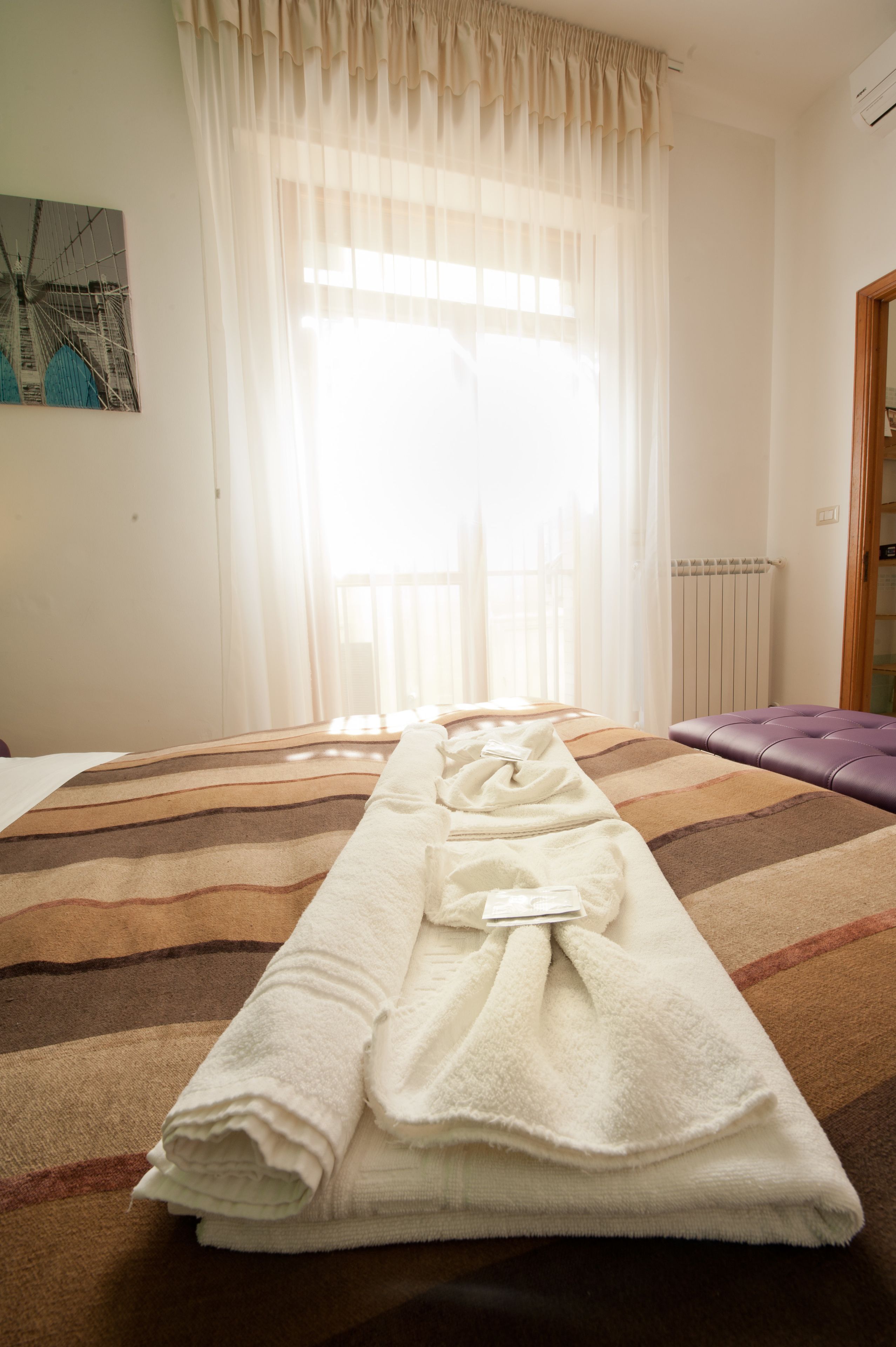 Une chambre d’hôtel avec un lit, une fenêtre ornée de rideaux légers, un radiateur et une serviette blanche pliée posée sur le lit.