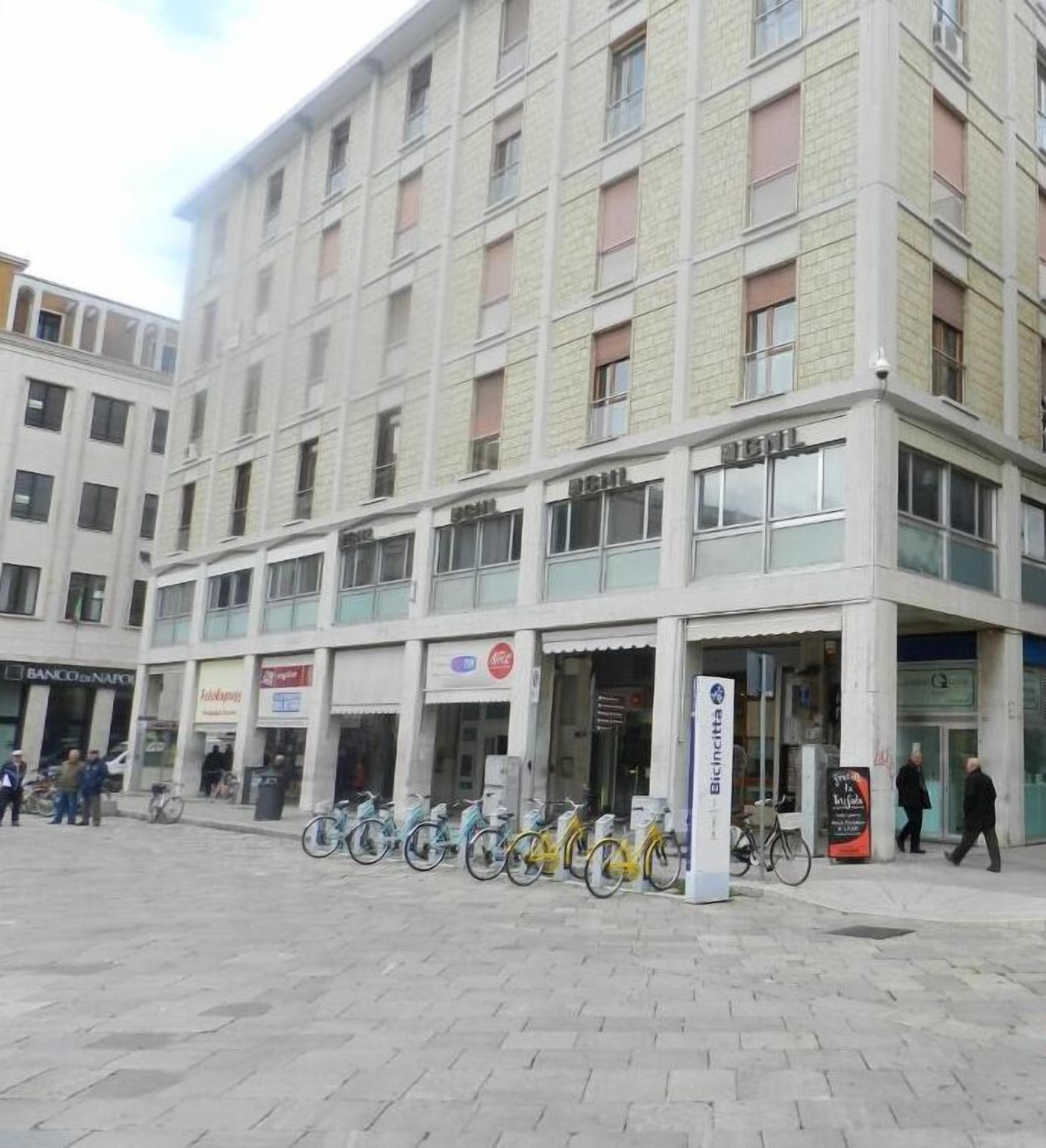 A multi-story building with a row of shops on the ground floor, a bike parking area, and a sign for Bicincitta.