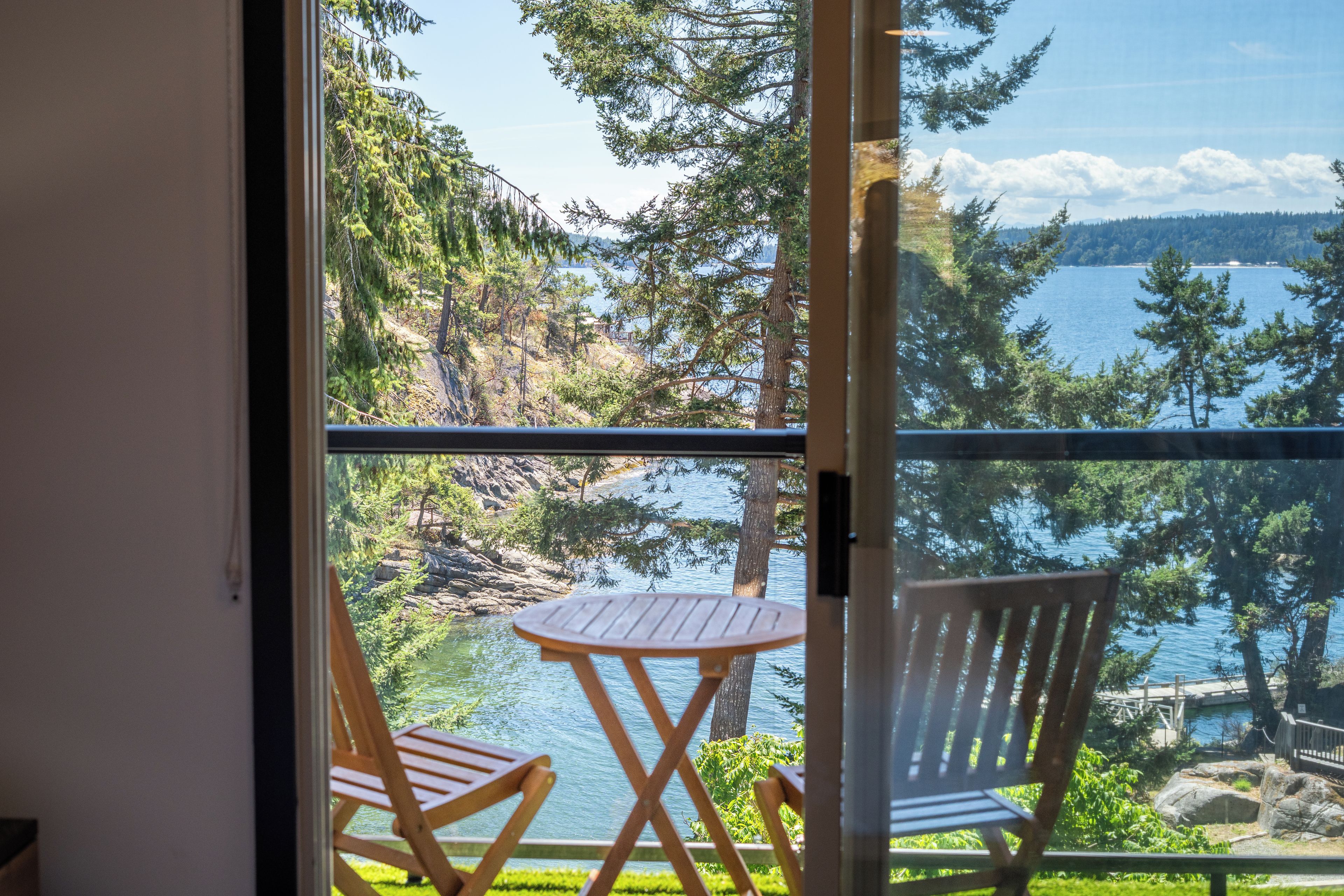 A balcony with two wooden chairs and a small round table, overlooking a lake with trees and a distant shoreline.
