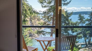 A balcony with two wooden chairs and a small round table, overlooking a lake with trees and a distant shoreline.