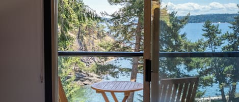 Un balcon avec deux chaises en bois et une petite table ronde, donnant sur un lac bordé d’arbres et dont on aperçoit la rive au loin.