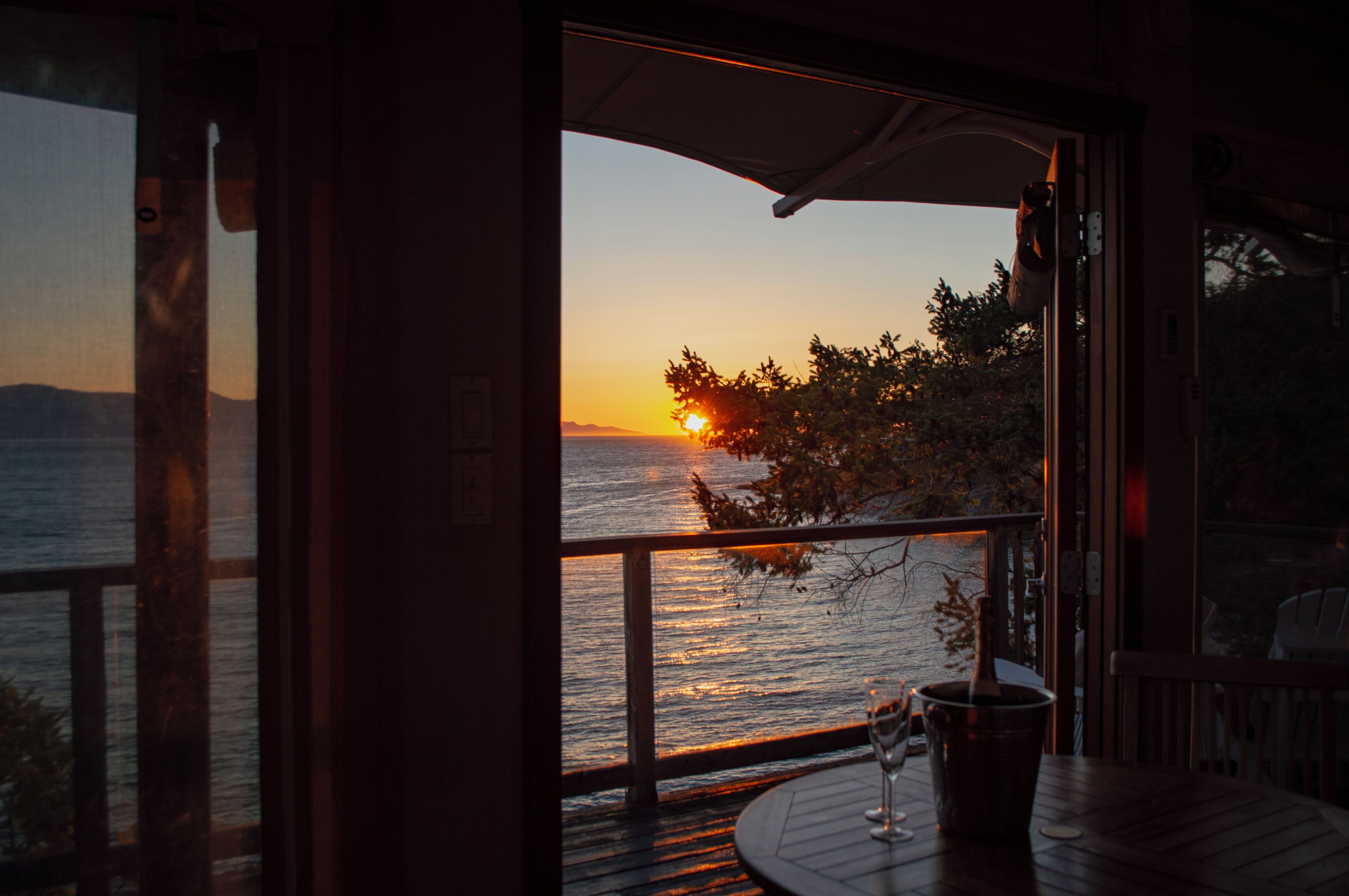 A sunset view from a room with a wooden deck, a glass door, and a table with a wine glass.