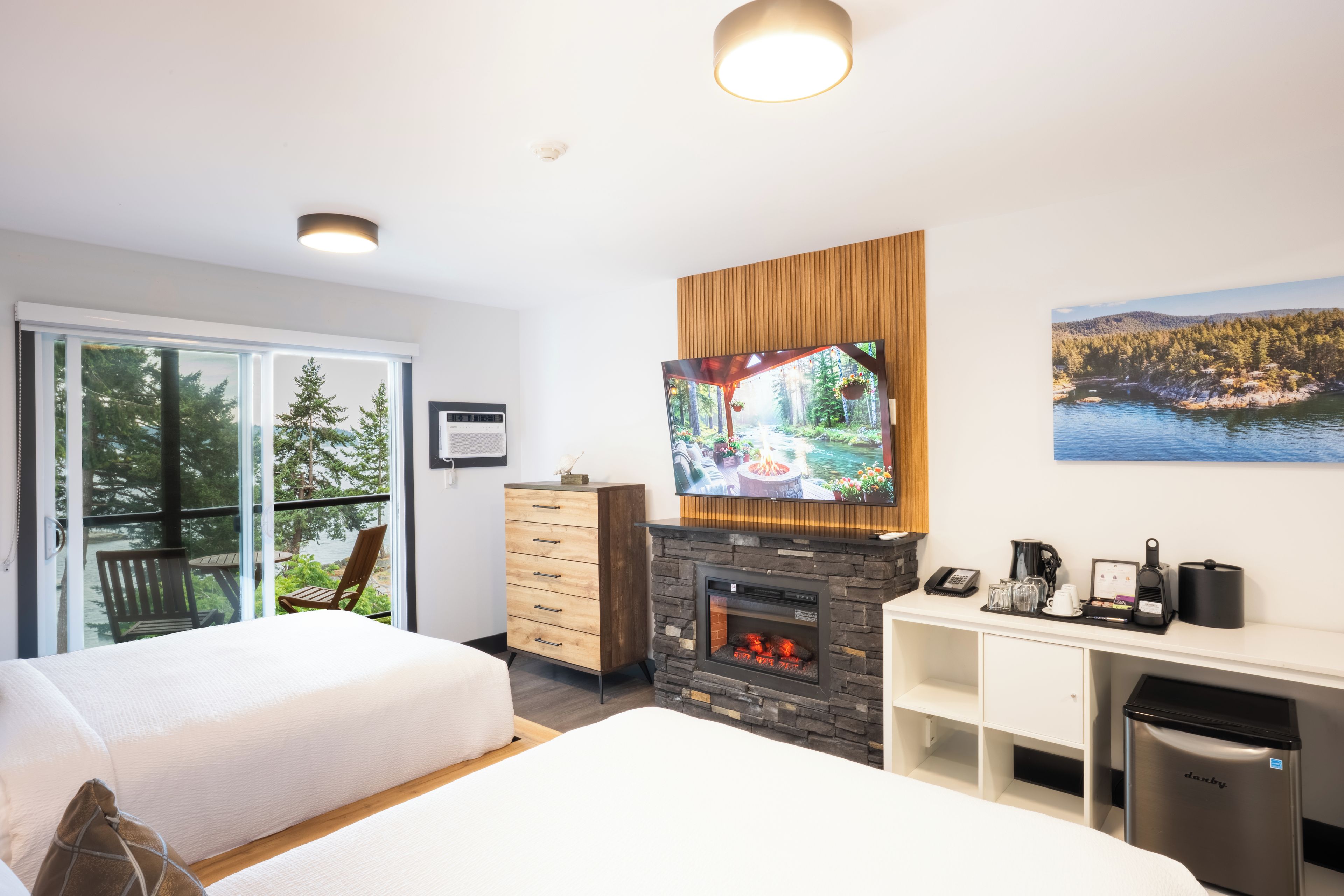 A modern hotel room with a large TV, a fireplace, and a balcony with a view of trees and water.