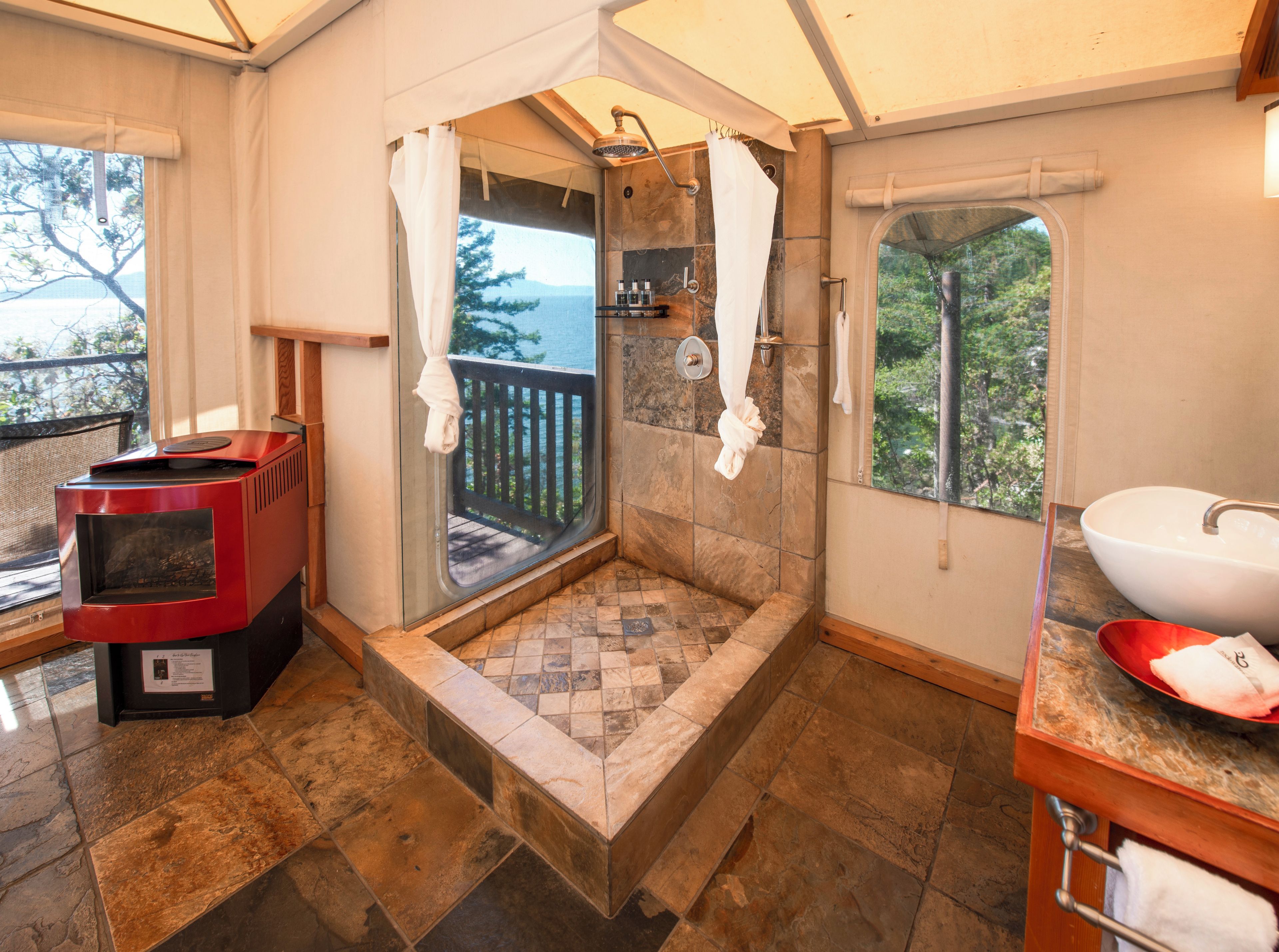 A bathroom with a stone-tiled floor, a red wood-burning stove, a large window with a view of the outdoors, a white sink, and a shower area.