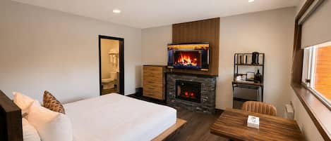 A modern hotel room with a bed, a fireplace, a dining table, and a small kitchen area.