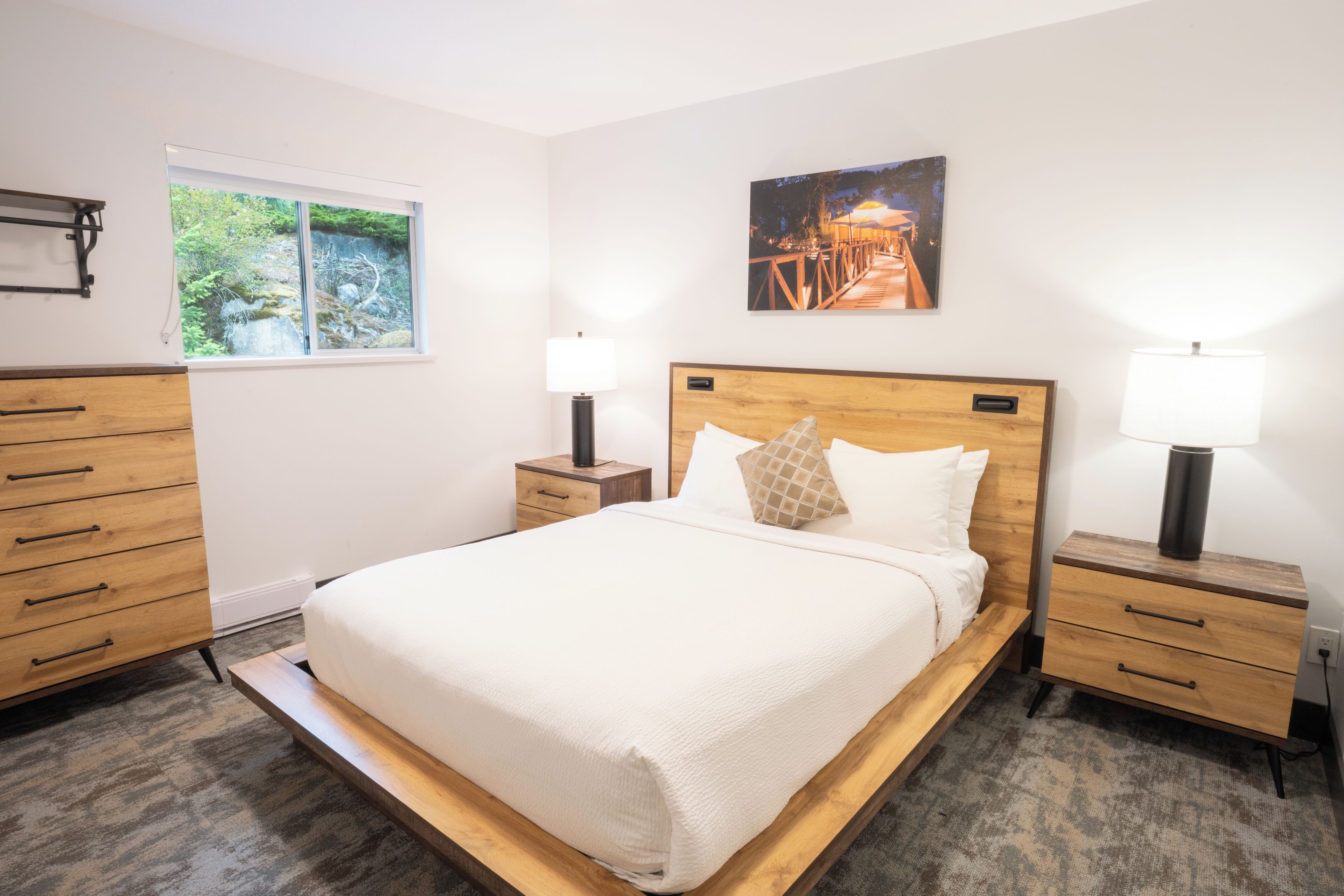 A bedroom with a bed, bedside tables, a nightstand, a window with a view of greenery, and a painting on the wall.