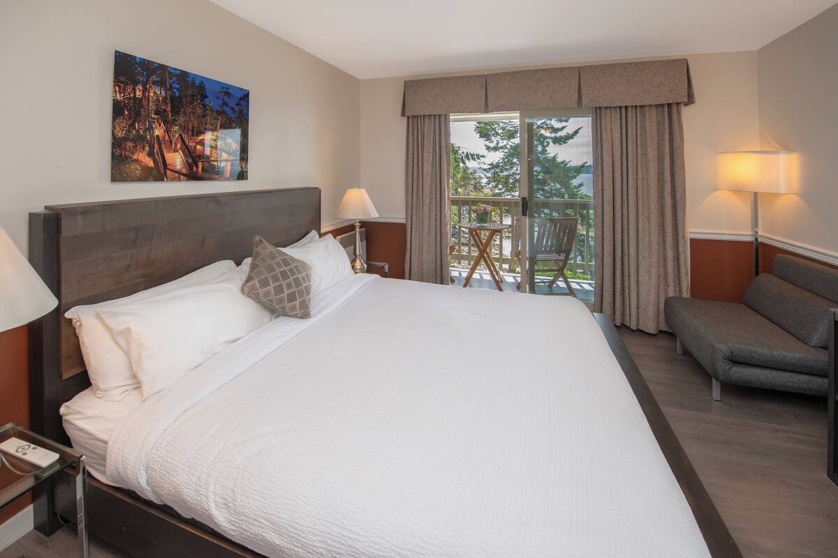 Quarto de hotel com uma cama grande, duas mesas de cabeceira com luminárias, um sofá, varanda com uma cadeira e um quadro na parede.