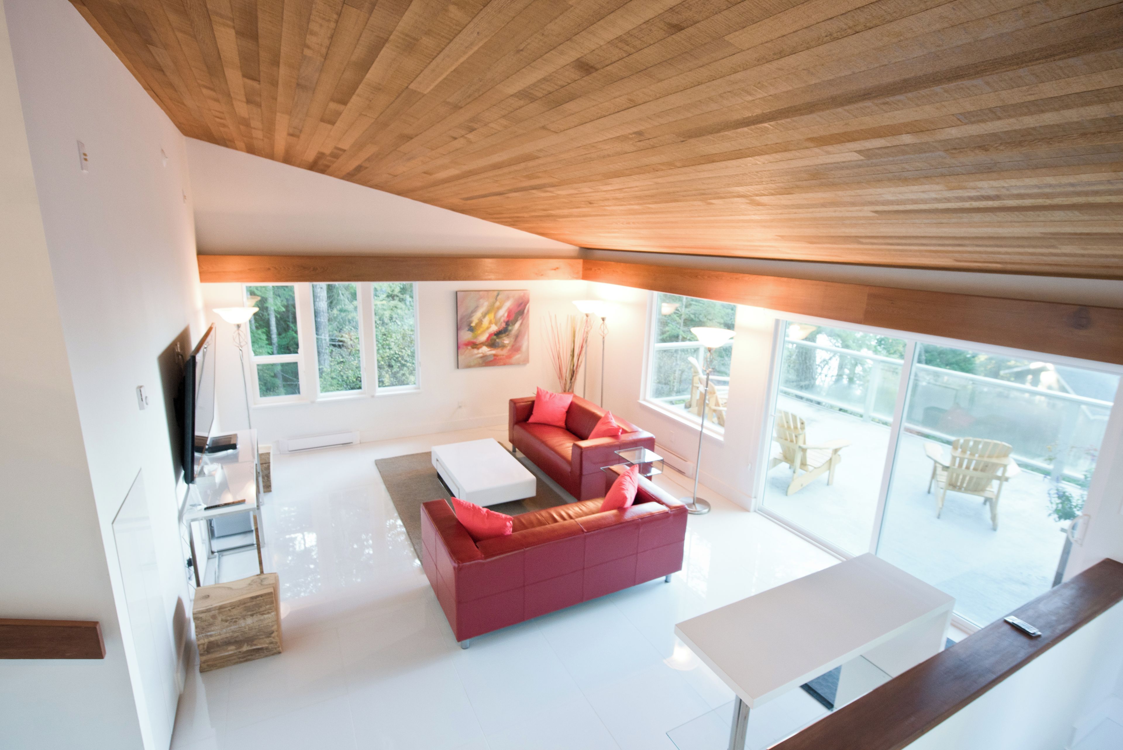 Una sala de estar moderna con un sofá rojo, una mesa de centro blanca y un televisor. Hay ventanas amplias y un balcón con mobiliario exterior.