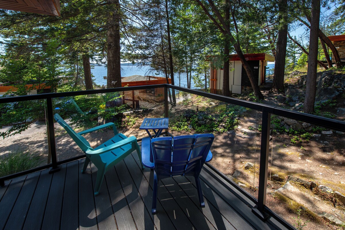 A wooden deck with two chairs and a table, overlooking a beach and a small cabin.