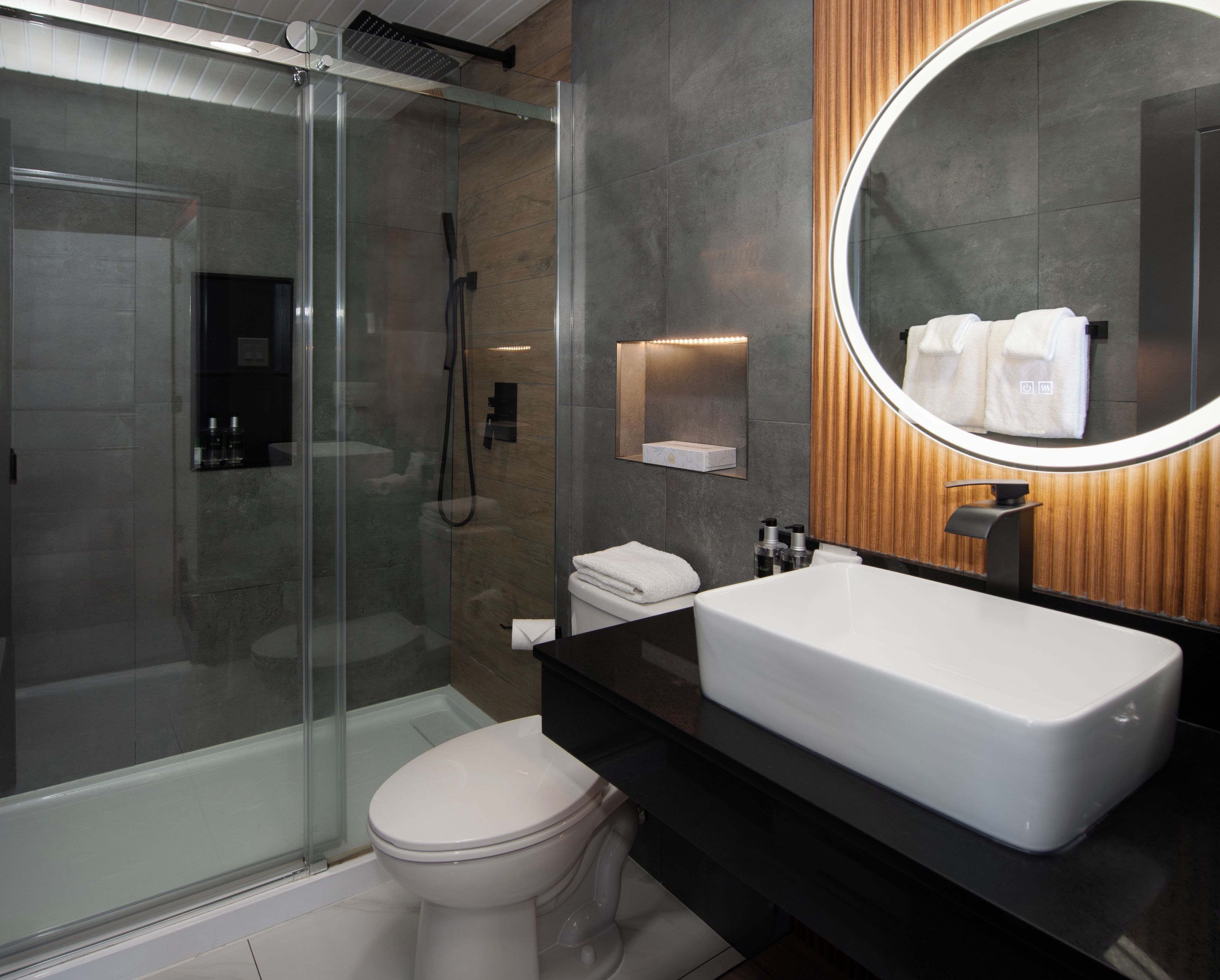 Un baño moderno con ducha de vidrio, inodoro blanco, lavamanos grande y espejo con marco de madera.