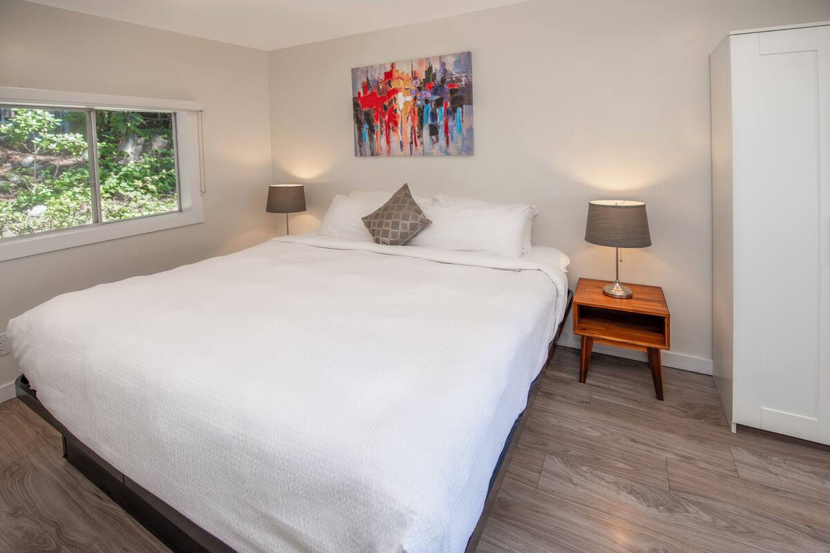 Quarto moderno com cama grande, duas mesas de cabeceira com luminárias, um quadro abstrato colorido na parede e uma janela com vista para o verde.