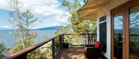 Une terrasse en bois offrant une vue sur la mer et les montagnes, une chaise en osier et une porte avec des panneaux de verre.
