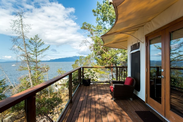 A wooden deck with a view of the sea and mountains, a wicker chair, and a door with glass panels.