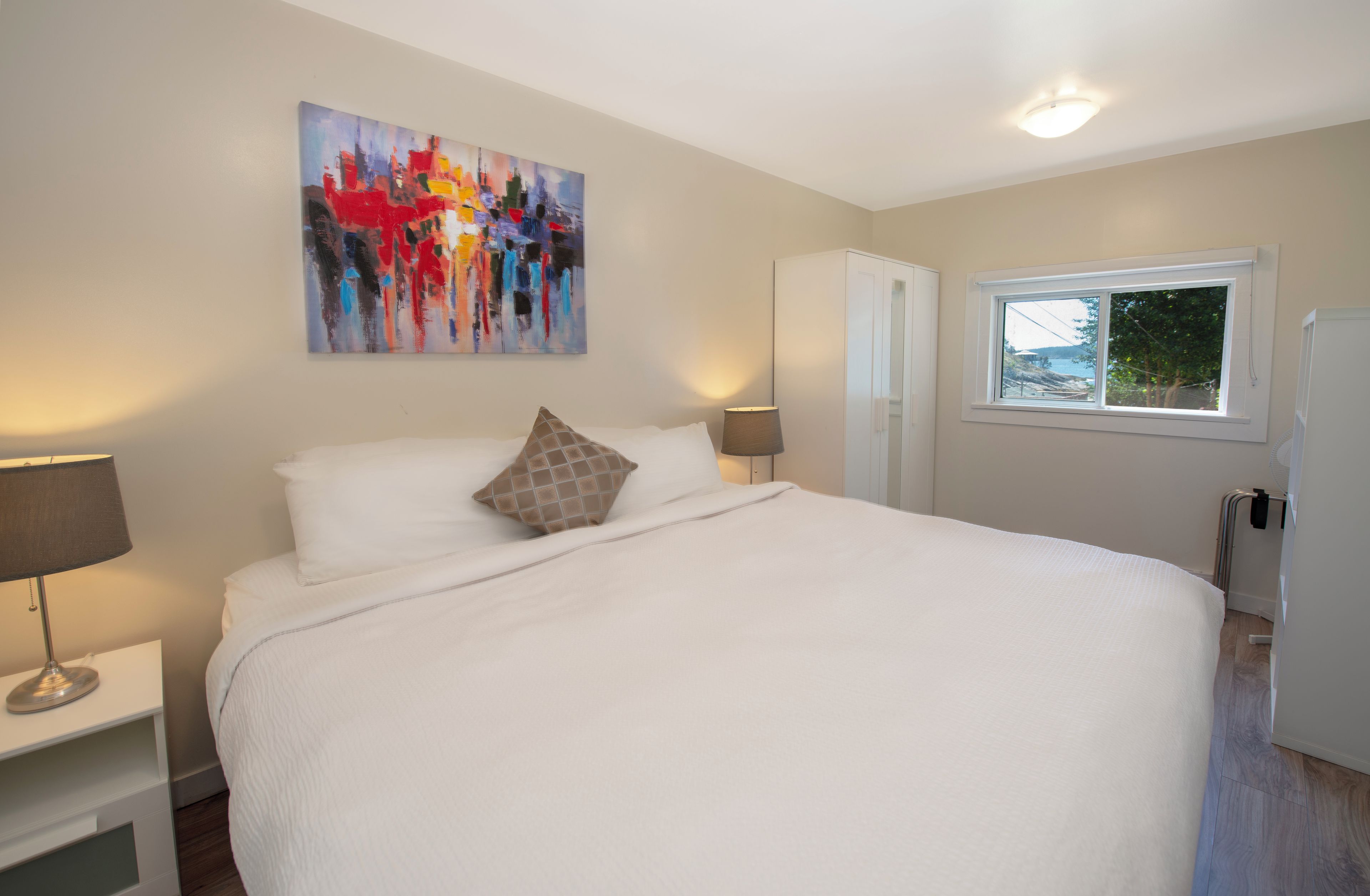 A bedroom with a large bed, a painting on the wall, a window, and a wardrobe.