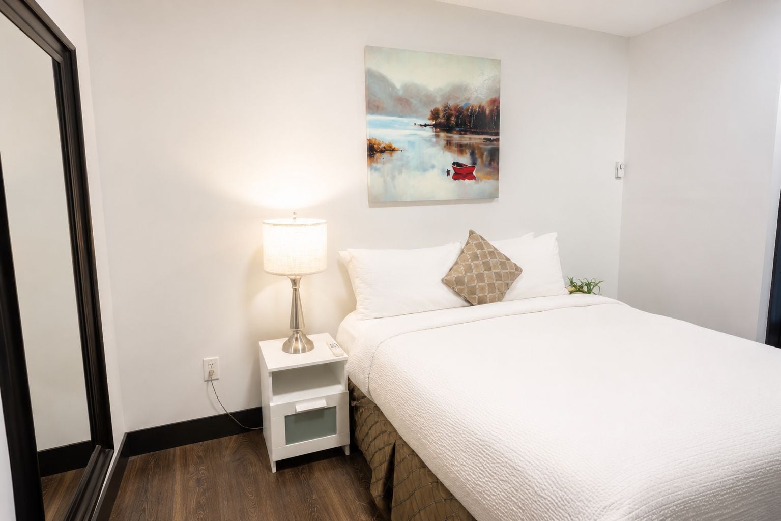 Una habitación de hotel con una cama, una mesita de noche con lámpara, un cuadro en la pared y una planta pequeña.