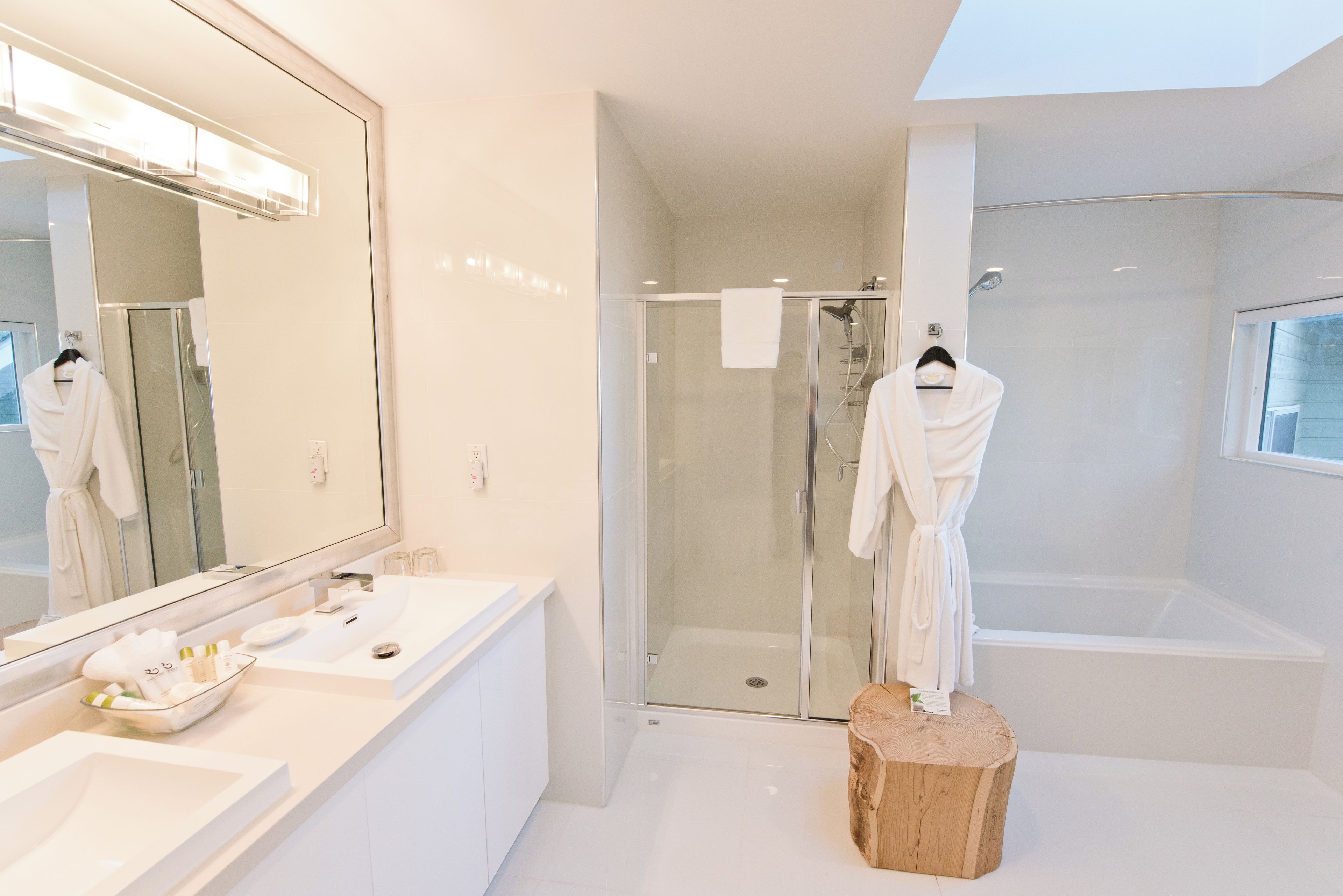 A modern bathroom with a large mirror, white fixtures, a glass shower enclosure, and a wooden stool.