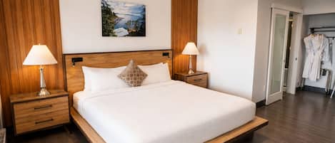 Una habitación de hotel con una cama grande, cabecera de madera, dos lámparas de noche, un cuadro en la pared y un perchero para bata.