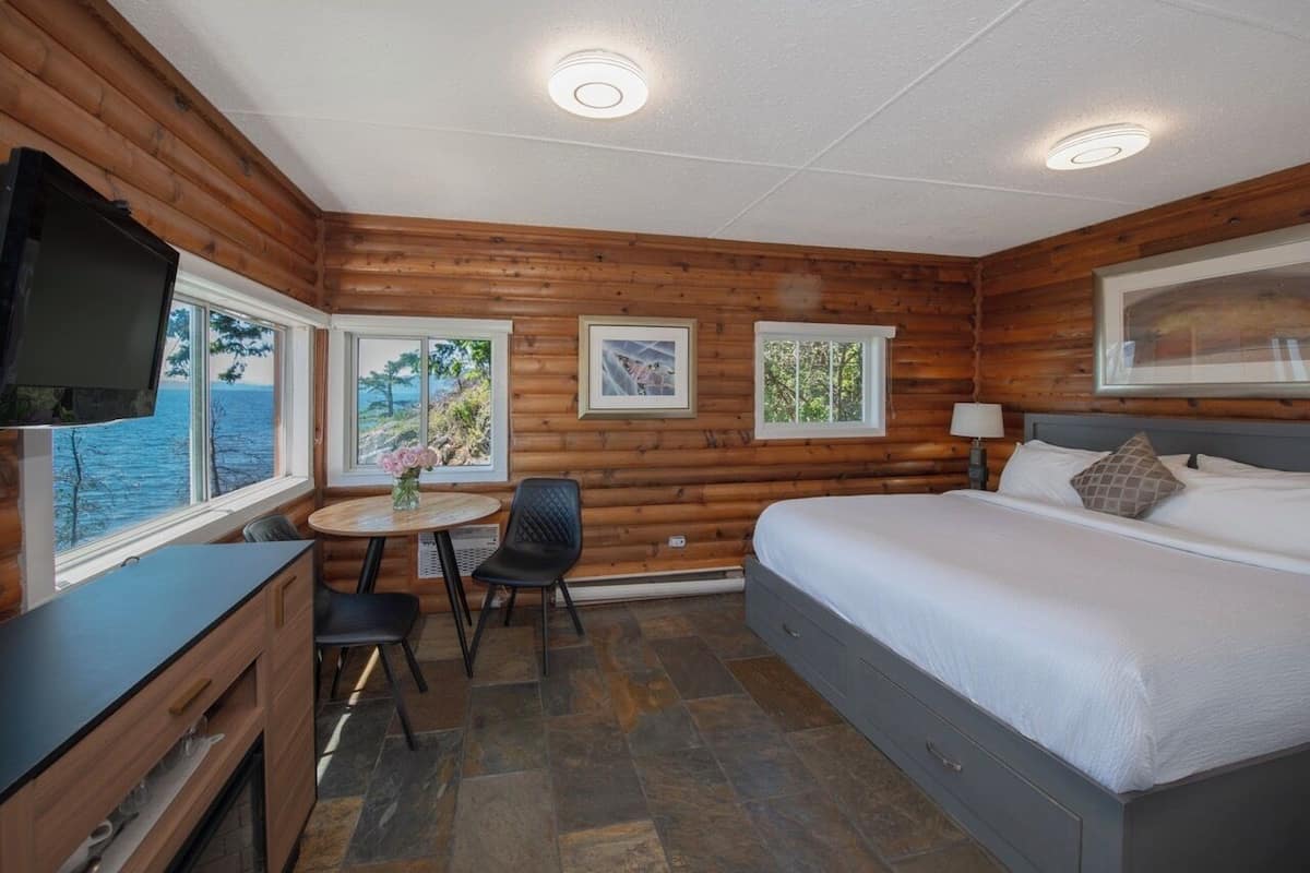 Quarto de cabana de tronco com cama, escrivaninha, cadeira, televisão e janela com vista para o mar.