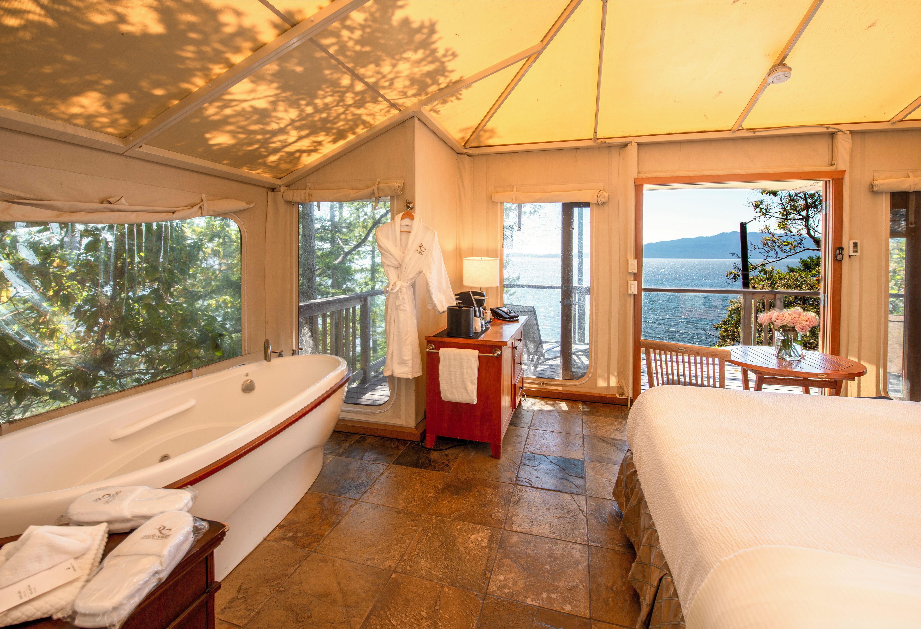 Una habitación con una bañera grande, una cama, una mesita con un jarrón de flores y vista al mar.
