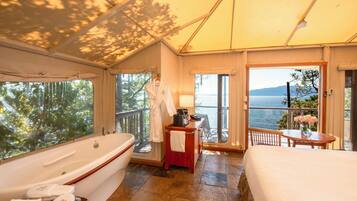 Una habitación con una bañera grande, una cama, una mesita con un jarrón de flores y vista al mar.