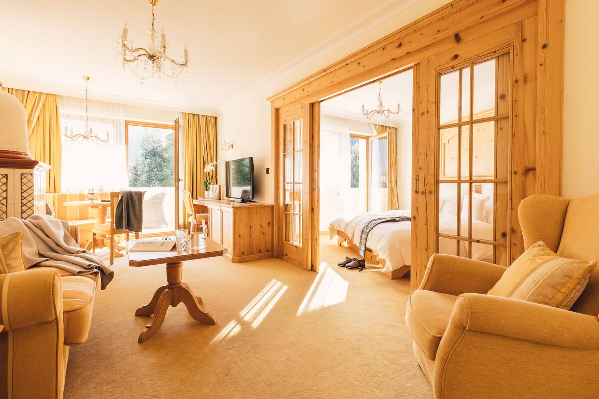 A spacious living room with a sofa, armchair, and a coffee table. There is a television on a wooden cabinet, and a bed is visible through an open door.