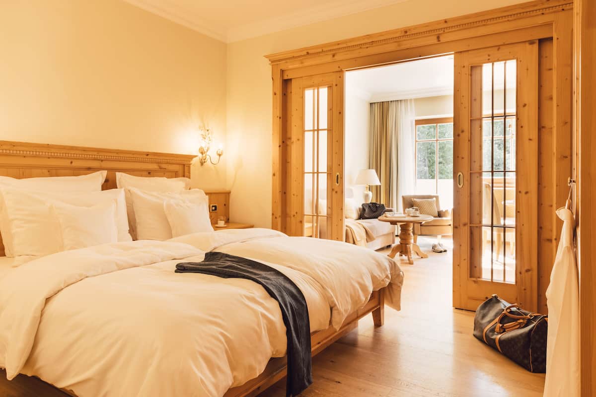 A hotel room with a large bed, wooden headboard, and a view through a wooden door to a sitting area with a table and chairs.