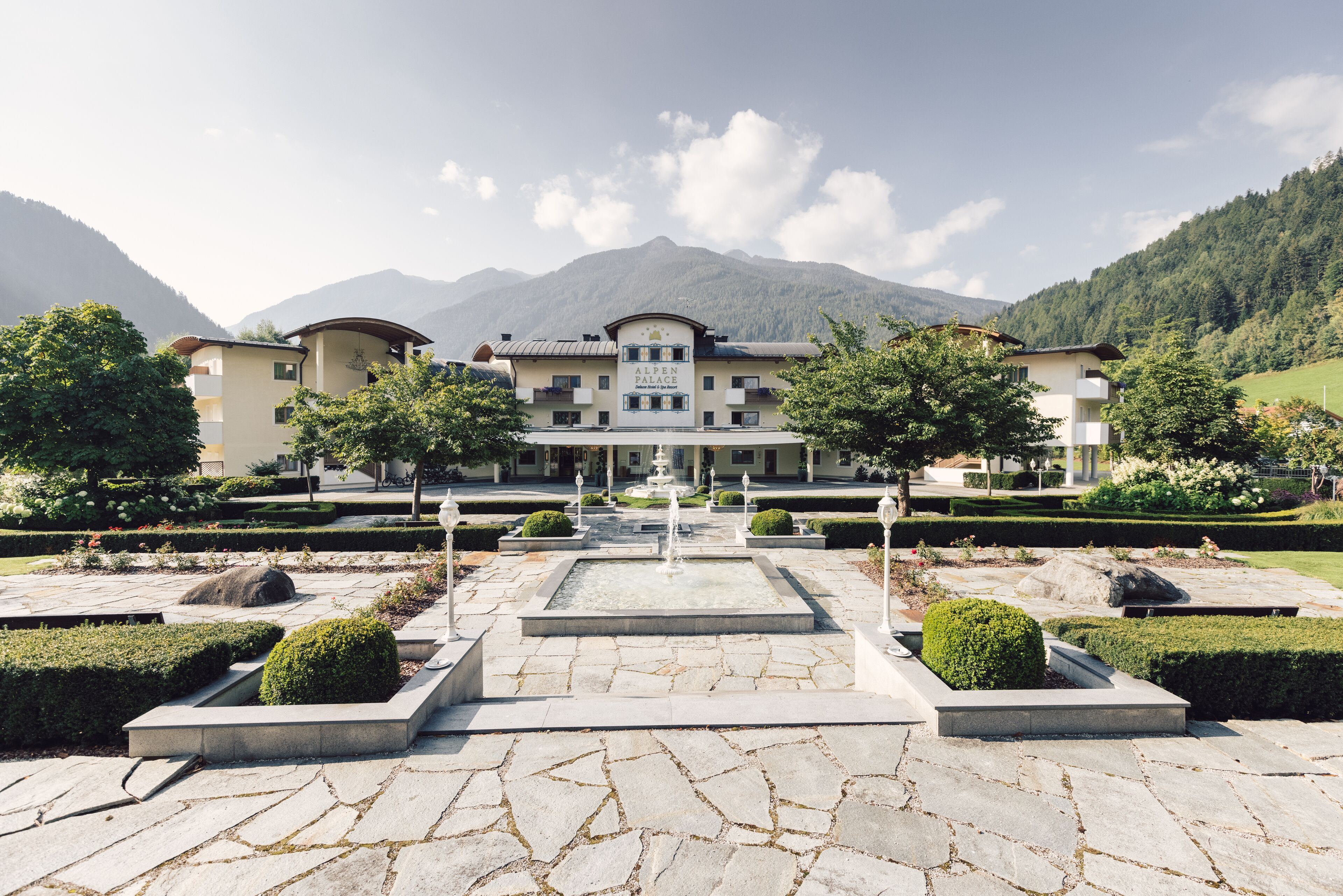 Un hotel grande de varios pisos con una fuente central, rodeado de jardines cuidados y montañas al fondo.