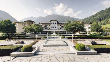 Un hotel grande de varios pisos con una fuente central, rodeado de jardines cuidados y montañas al fondo.