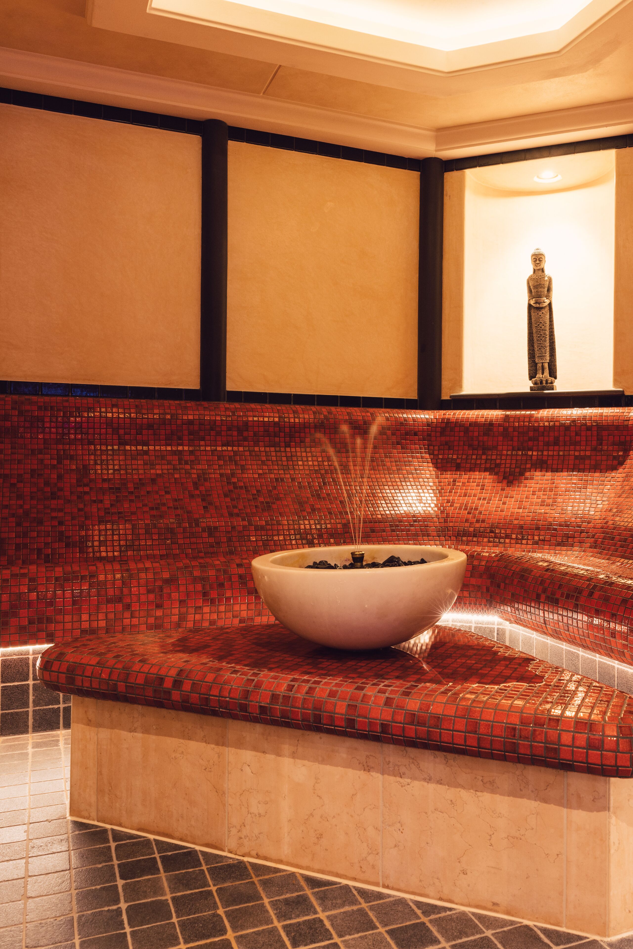 A spa area with a mosaic-tiled bench, a white bowl with black stones, and a statue on a wall.