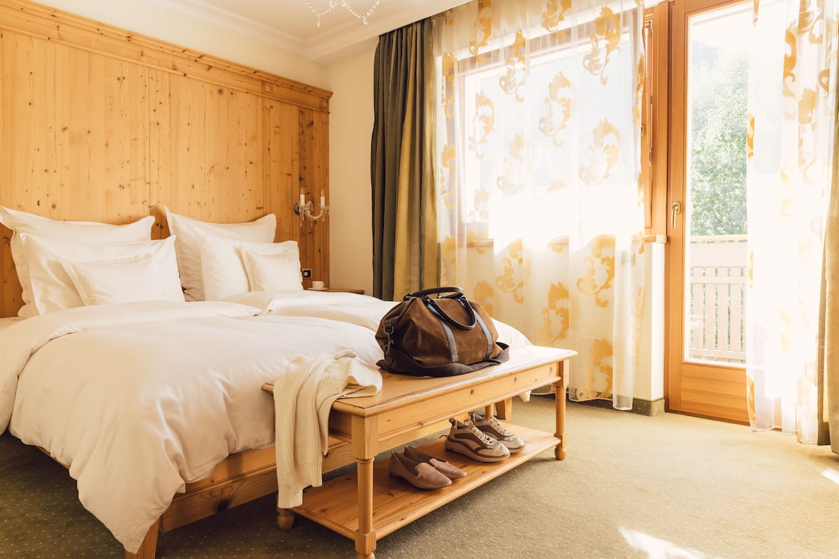 A neatly made bed with pillows, a wooden bedside table with a brown bag and shoes, and a window with curtains.