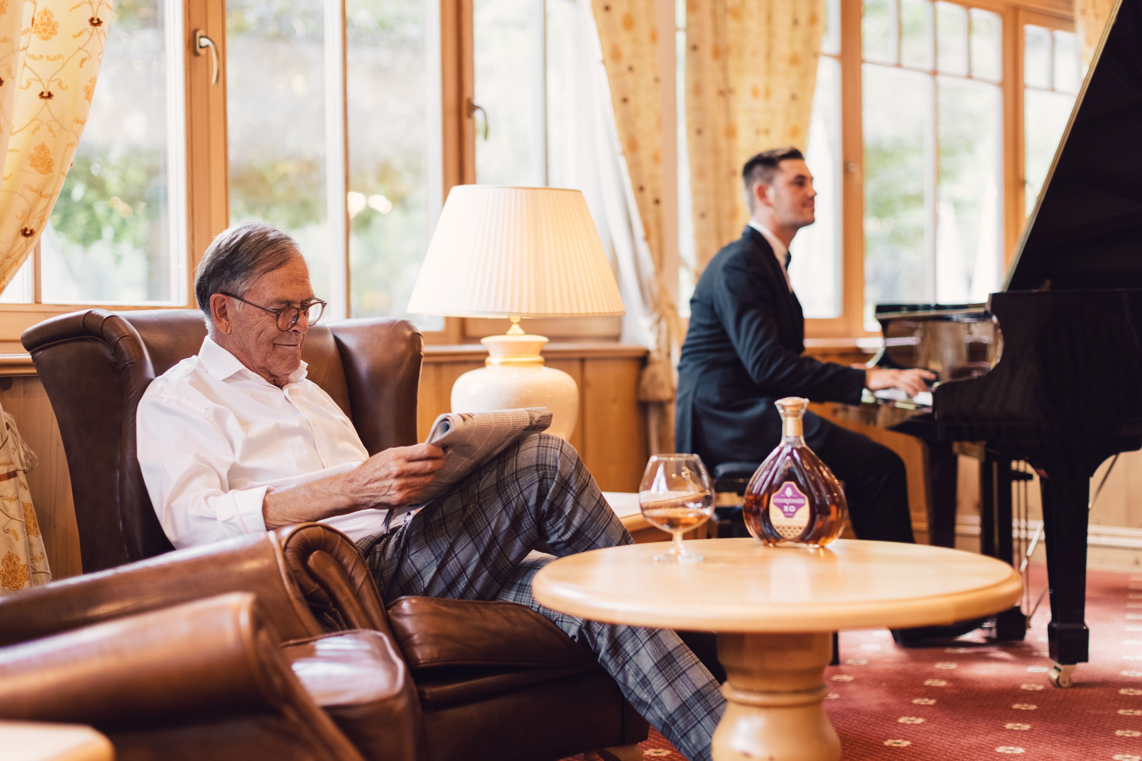 Un hombre leyendo en un sillón de cuero, una mesa con una botella y un vaso, y un piano de cola al fondo.
