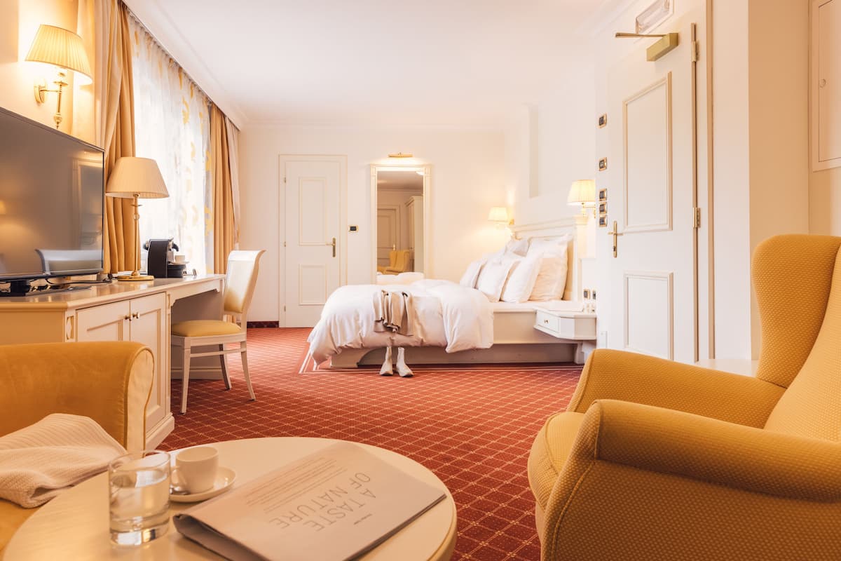 A hotel room with a bed, a desk with a lamp, a chair, and a table with a glass of water and a menu.