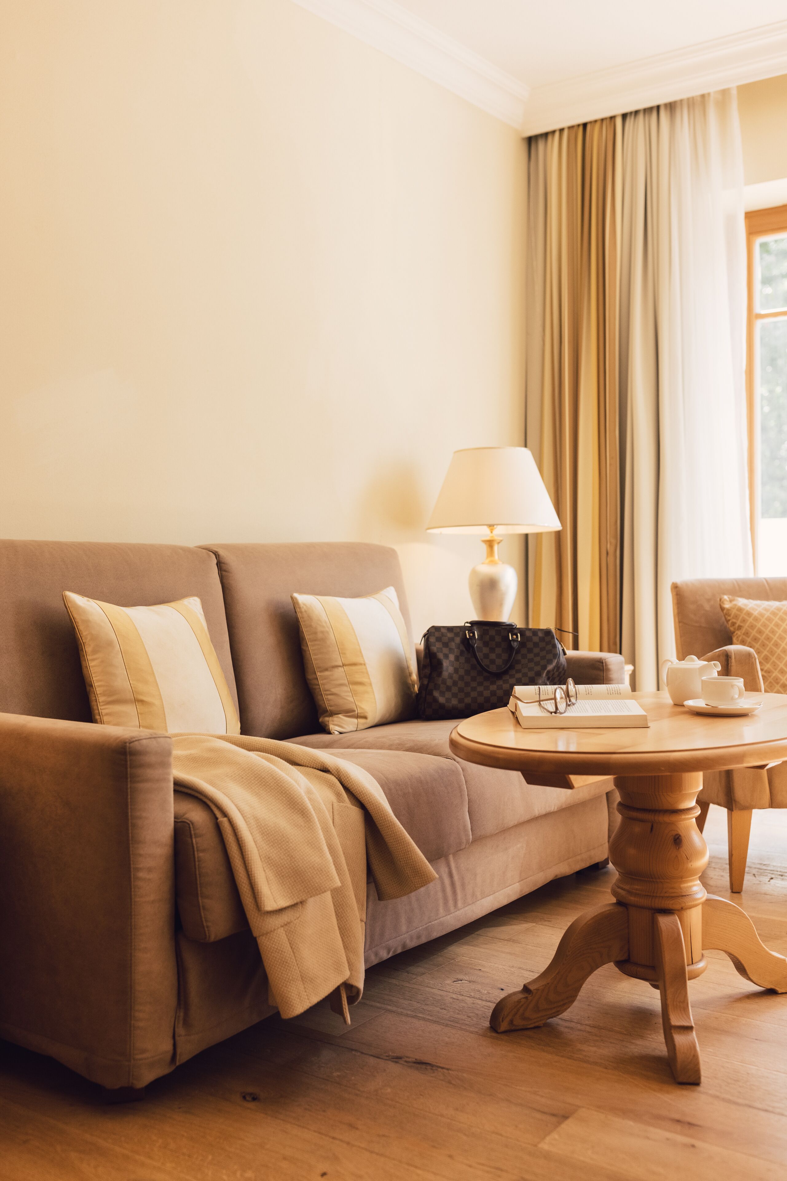 Habitación de hotel con un sofá color beige, una mesa de centro de madera, una lámpara, un bolso, una taza y un libro.