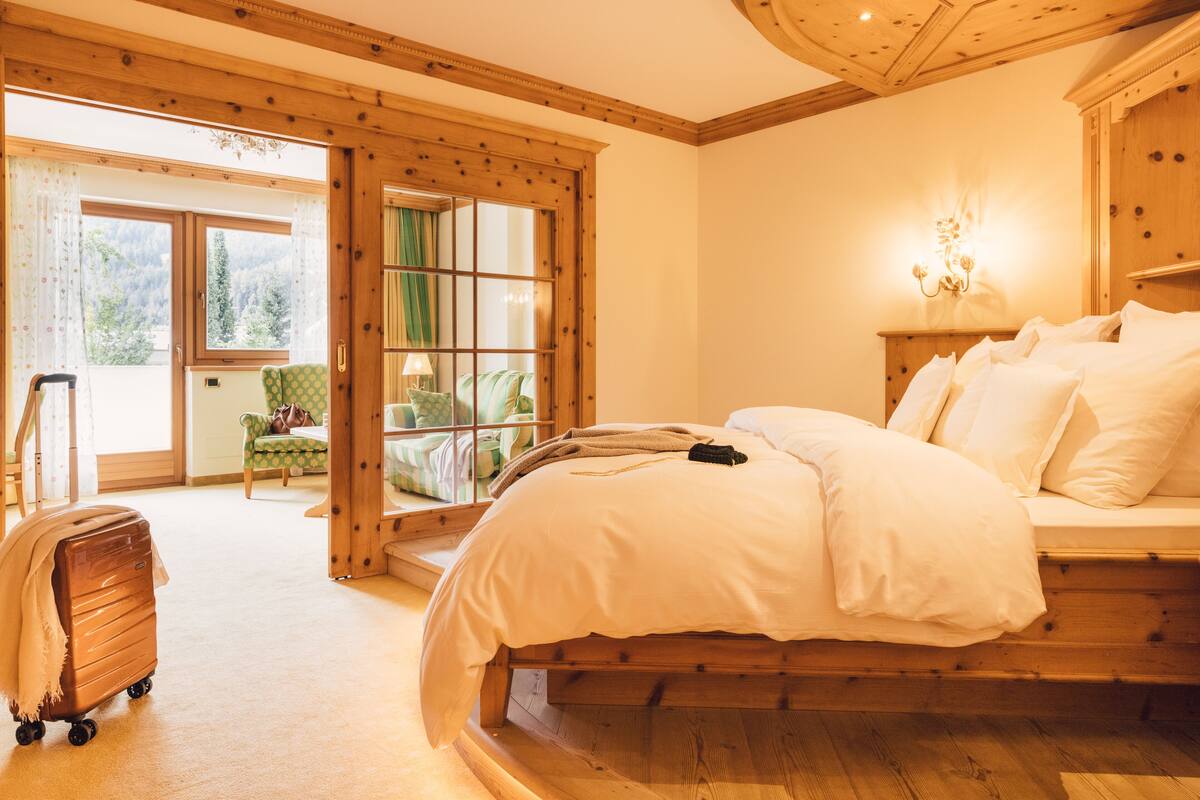 A bedroom with a large bed, wooden floor, a suitcase, and a view of a balcony with a chair and a table.