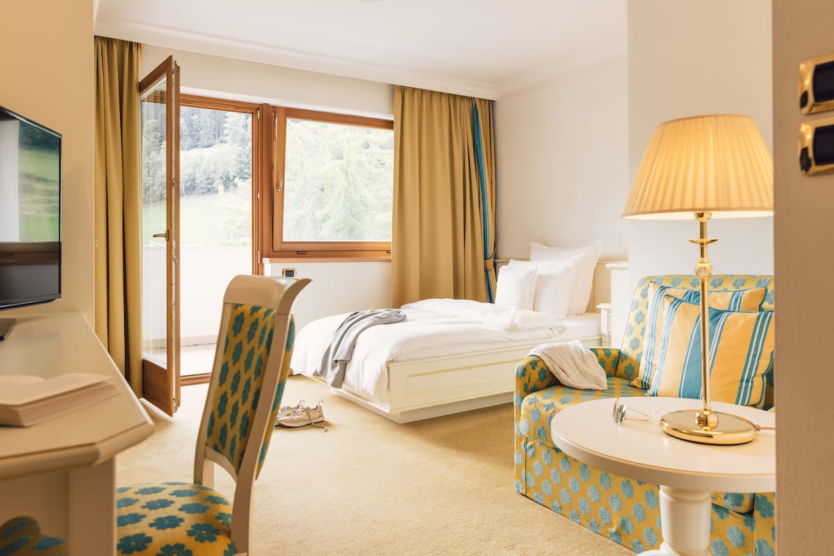 A hotel room with a bed, a desk with a TV, a chair, a table with a lamp, and a view of the outdoors.
