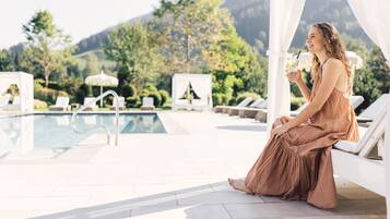 A woman sitting by a poolside, holding a drink, with lounge chairs and umbrellas in the background.