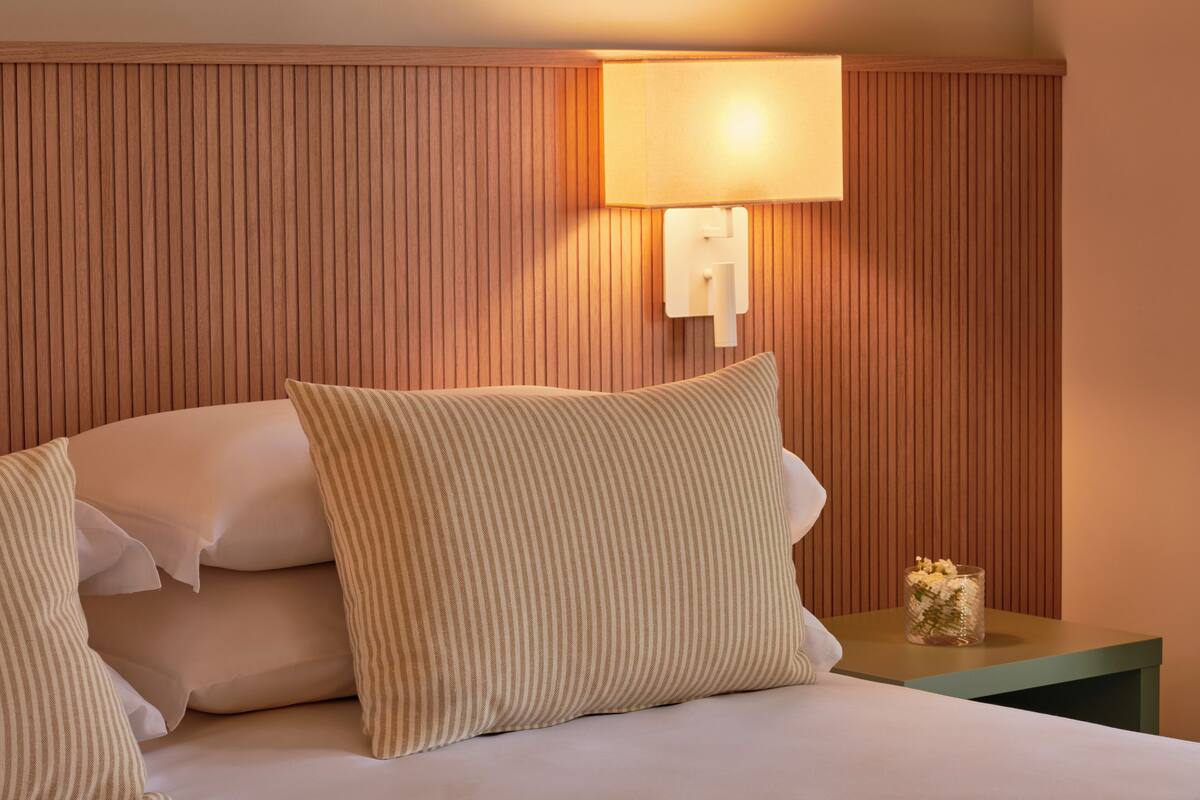 A neatly made bed with white linens and striped pillows, a bedside table with a glass vase of flowers, and a wall-mounted lamp.