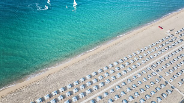 An aerial view of a beach with rows of beach umbrellas and a few sailboats in the water. - Serenè Resort (Cutro)