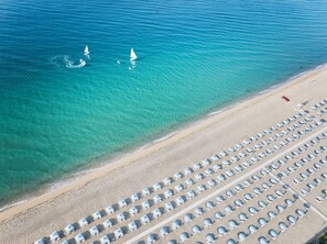 An aerial view of a beach with rows of beach umbrellas and a few sailboats in the water.